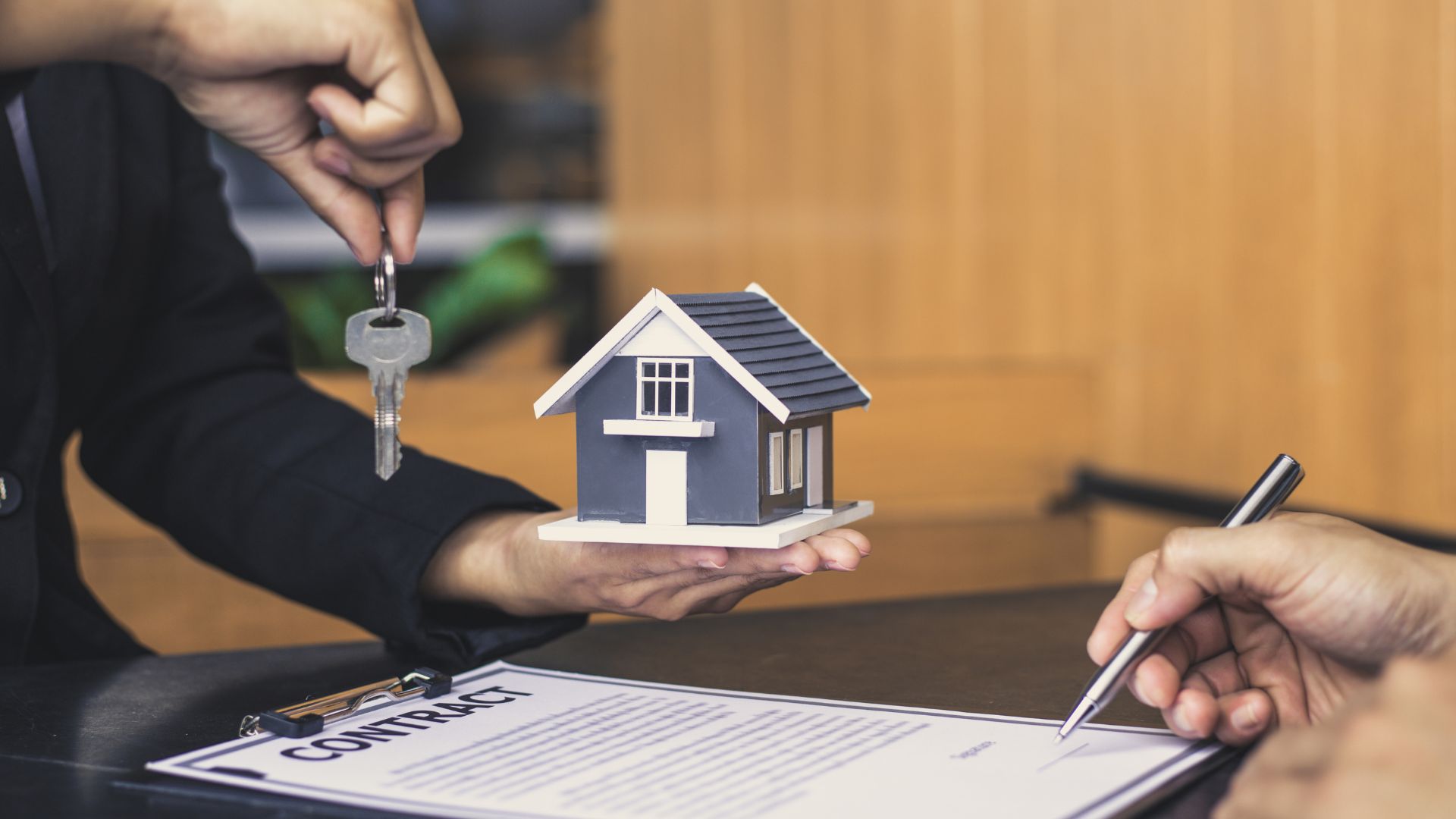 Hands exchanging house keys and miniature home over real estate contract